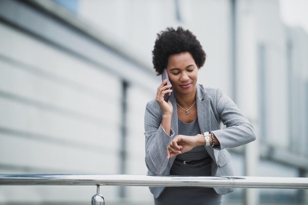 Retrato de una mujer de negocios negra sonriente hablando por teléfono inteligente y mirando un reloj de mano frente a un edificio corporativo.