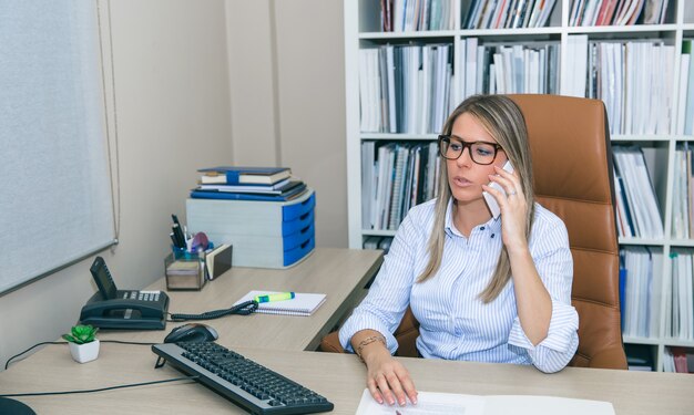 Retrato de mujer de negocios rubia hablando con smartphone sentado en la oficina