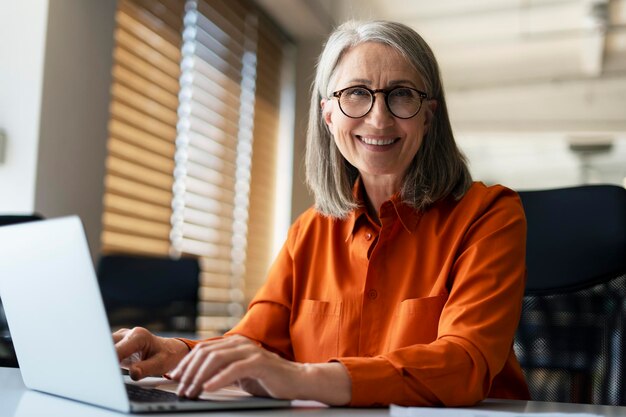 Retrato de una mujer de negocios senior sonriente y segura que usa anteojos elegantes usando una computadora portátil trabajando en línea en una oficina moderna Un financiero exitoso escribiendo en el teclado sentado en el lugar de trabajo