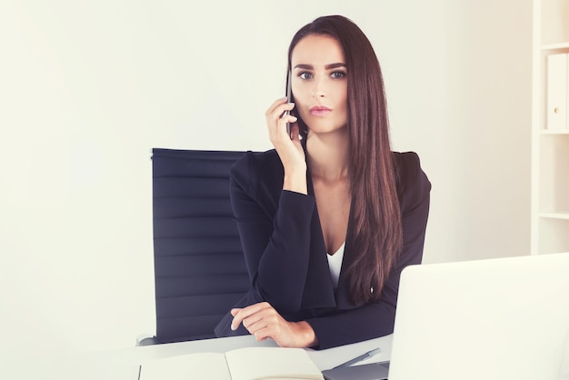 Retrato de una mujer de negocios seria con cabello castaño largo sentada en su lugar de trabajo y hablando por teléfono. imagen tonificada