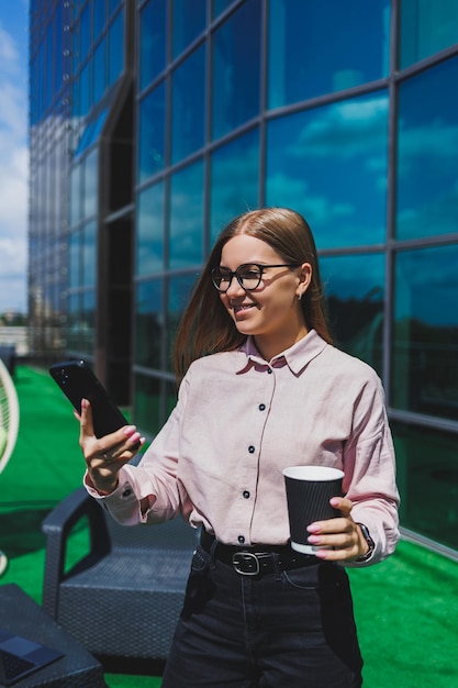 Retrato de mujer de negocios sonriente leyendo sms en el teléfono inteligente durante la pausa para el café en el balcón de la oficina Terraza moderna en la oficina para relajarse