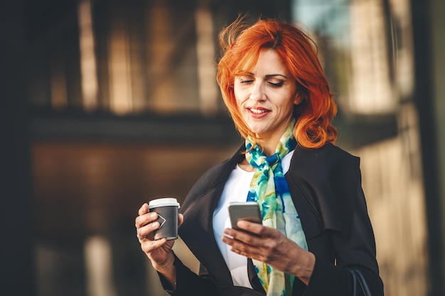 Retrato de una mujer de negocios sonriente navegando en un smartphone en un descanso para tomar café en el distrito de oficinas en el día ventoso. Ella está mirando el teléfono inteligente con el pelo volador.