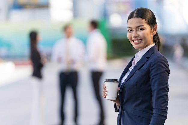 Retrato de mujer de negocios sonriente sosteniendo una taza de café mientras está de pie frente a modernos edificios de oficinas