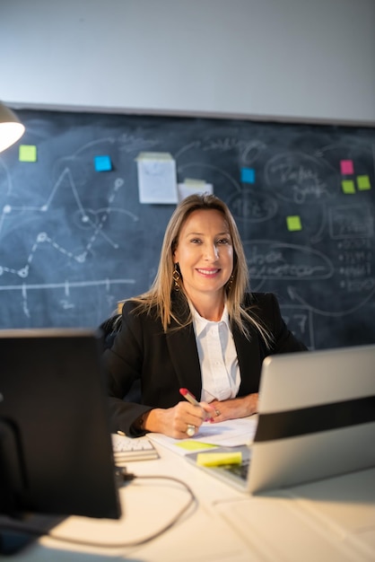 Retrato de mujer de negocios trabajando por la noche. Mujer feliz trabajando en la mesa en la oficina, haciendo análisis financieros anotando resultados con bolígrafo y mirando la cámara. Concepto de finanzas y crecimiento empresarial