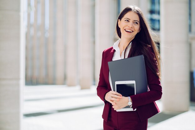 El retrato de la mujer de negocios se vistió en el traje marrón, sostiene la tableta y la carpeta, sonriendo ampliamente al aire libre. Concepto de gente de negocios