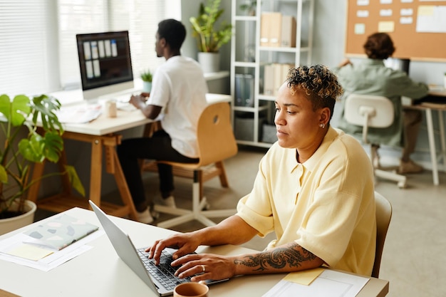 Retrato de una mujer negra tatuada que usa una computadora portátil en la oficina mientras trabaja con un equipo en una copia de fondo