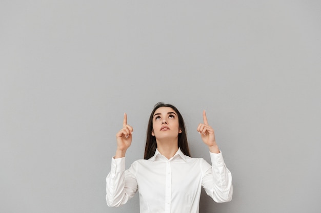 Retrato de mujer de oficina inteligente con cabello largo castaño en camisa blanca mirando hacia arriba y apuntando con el dedo hacia arriba en copyspace, aislado sobre pared gris