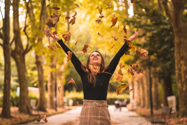 Retrato de una mujer en otoño en un bosque con hojas marrones al atardecer en un parque de la ciudad tirando hojas