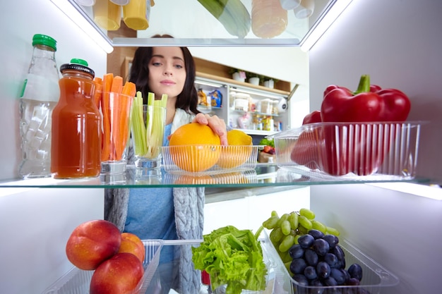 Retrato de mujer parada cerca de una nevera abierta llena de alimentos saludables, verduras y frutas.