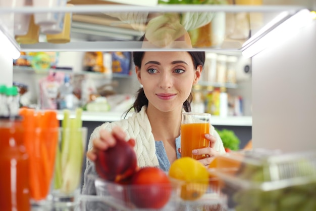 Retrato de mujer parada cerca de una nevera abierta llena de frutas y verduras saludables