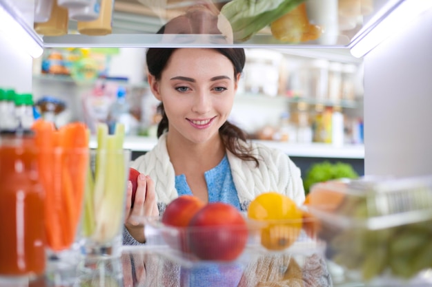 Retrato de mujer parada cerca de una nevera abierta llena de frutas y verduras saludables