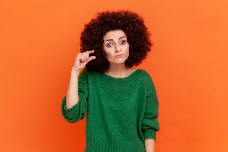 Foto retrato de mujer con peinado afro con suéter de estilo informal verde que muestra un gesto de tamaño pequeño con los dedos mirando a la cámara con decepción toma de estudio interior aislada sobre fondo naranja