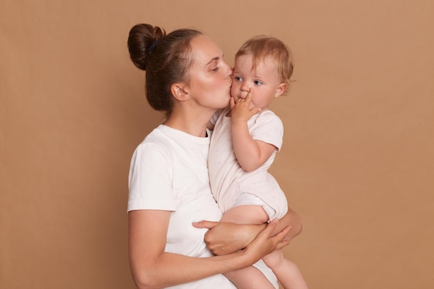 Retrato de mujer con peinado de moño en camiseta blanca de pie con su madre besando a su hija pequeña expresando una pose suave aislada sobre fondo marrón