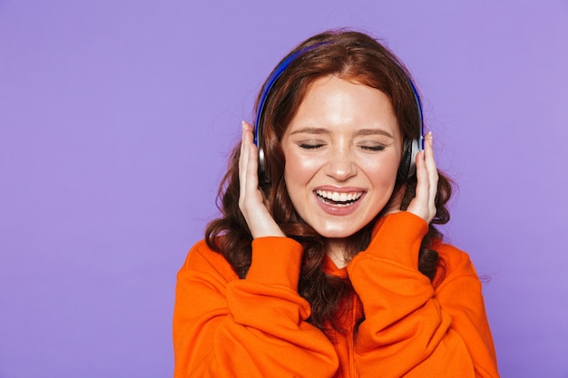 Retrato de una mujer pelirroja bastante joven de pie sobre violeta, escuchando música con auriculares