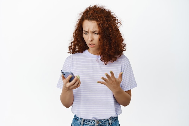 Foto retrato de mujer pelirroja confundida que no puede entender cómo usar la aplicación, mala experiencia de usuario con la aplicación de teléfono inteligente, mirando el teléfono desconcertado y molesto en blanco