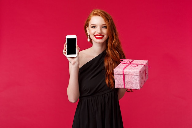 Retrato de mujer pelirroja elegante y con estilo que muestra la pantalla del teléfono móvil y una caja de regalo rosa, recomiende la tienda o el servicio de entrega para comprar regalos para su niña, sonriente, en una pared roja