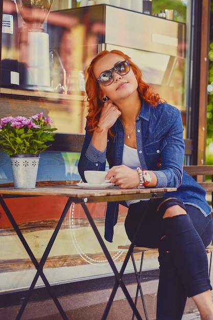 Retrato de mujer pelirroja con gafas de sol, bebe café en una cafetería en una calle.