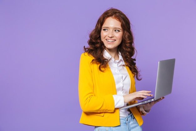 Retrato de una mujer pelirroja joven muy sonriente de pie sobre violeta, trabajando en equipo portátil