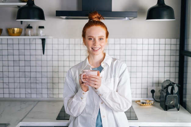 Retrato de mujer pelirroja sonriente sosteniendo una taza de bebida caliente en las manos en la mañana en la cocina