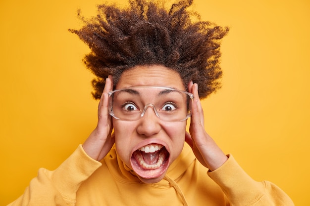 Retrato de mujer de pelo rizado conmocionada emocionalmente agarra la cara y grita en voz alta mantiene la boca abierta