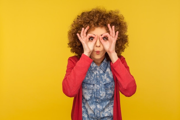 Retrato de una mujer con el pelo rizado vestida de forma informal, mirando a través del gesto de los binoculares y expresando sorpresa, ampliando la visión, explorando la distancia. tiro de estudio interior aislado sobre fondo amarillo