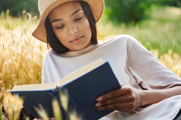 Retrato de mujer de pensamiento vistiendo piercing de labios y sombrero de paja libro de lectura mientras está acostado sobre el césped en el parque verde
