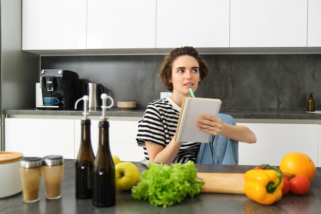 Retrato de una mujer pensante con un cuaderno de cocina escribiendo los ingredientes de la receta decidiendo una comida