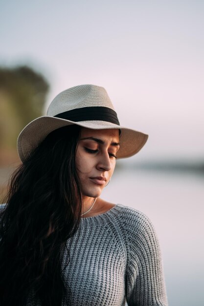 Retrato de una mujer pensativa y seria con los ojos cerrados con un sombrero junto a un lago