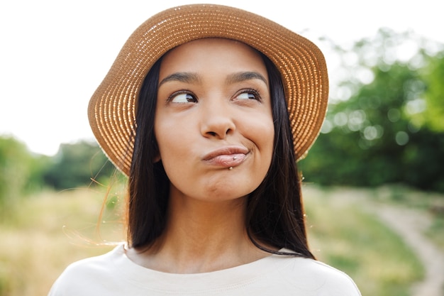 Retrato de mujer pensativa con sombrero de paja y piercing en el labio mirando a un lado mientras camina en el parque verde