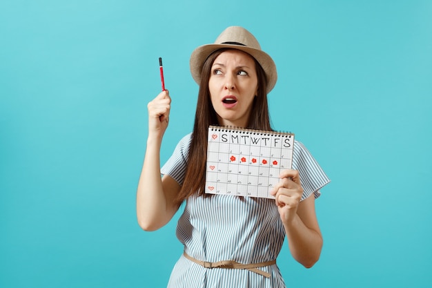 Retrato de mujer pensativa en vestido azul, sombrero con lápiz rojo, calendario de períodos femeninos para comprobar los días de menstruación aislados sobre fondo azul. Atención médica, concepto ginecológico. Copia espacio