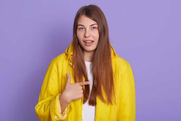 Retrato de mujer pidiéndote hablar conmigo, te refieres a mí, mujer bonita apuntando a sí misma aislada sobre fondo lila, vestida con chaqueta amarilla casual.