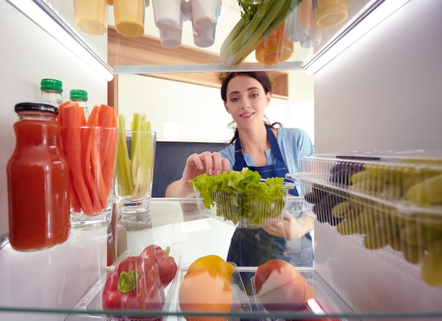 Retrato de mujer de pie cerca de una nevera abierta llena de alimentos saludables, verduras y frutas, retrato de