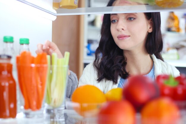 Retrato de mujer de pie cerca de una nevera abierta llena de alimentos saludables, verduras y frutas