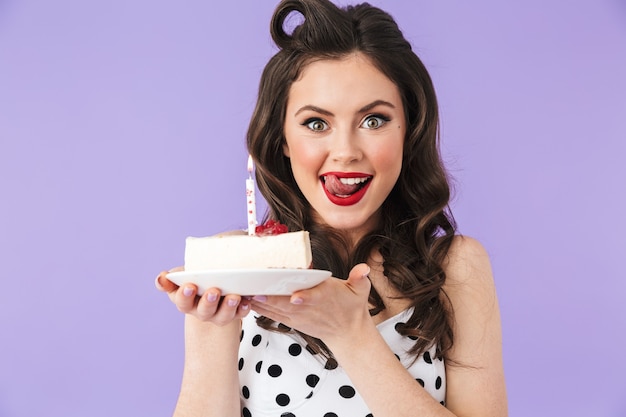 Retrato de mujer pin-up americana en vestido de lunares vintage celebrando y sosteniendo tarta de queso de cumpleaños con vela aislada sobre pared violeta