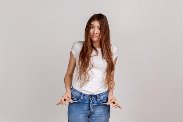 Retrato de una mujer pobre disgustada con el pelo oscuro que saca los bolsillos vacíos preocupados por las deudas sin dinero para vivir usando una camiseta blanca Foto de estudio interior aislada en un fondo gris