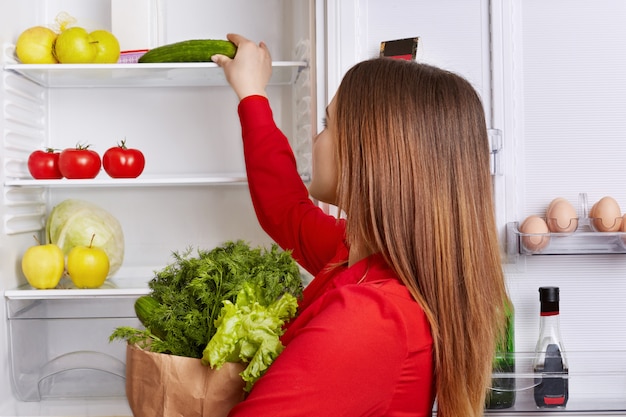 Retrato de mujer pone verduras en los estantes del refrigerador