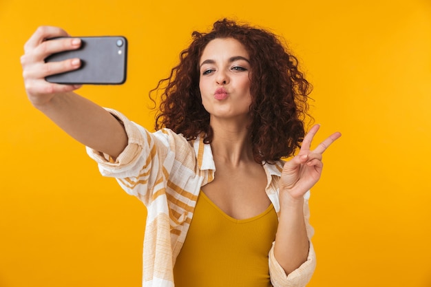 Foto retrato de mujer positiva de 20 años con cabello rizado sonriendo y tomando fotos selfie en smartphone, aislado en amarillo