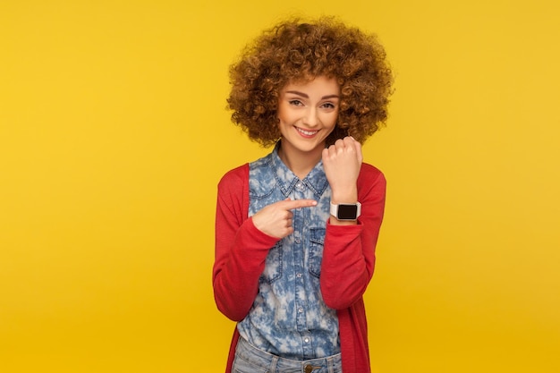 Retrato de una mujer positiva y alegre con el pelo rizado que muestra un reloj de pulsera y mira alegremente a la cámara, recordando la hora tardía, pidiendo prisa. tiro de estudio interior aislado sobre fondo amarillo