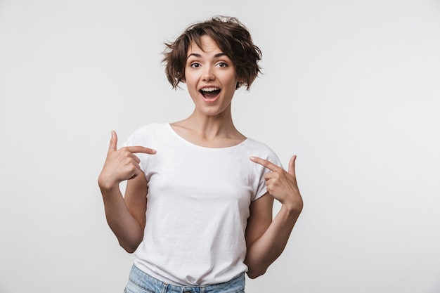 Retrato de mujer positiva con cabello castaño corto en camiseta básica regocijándose y señalando con el dedo a sí misma aislada sobre pared blanca