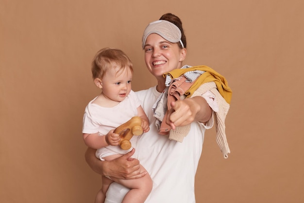 Retrato de mujer positiva feliz con camiseta blanca y máscara para dormir madre con hija sosteniendo ropa en el hombro señalando con el dedo a la cámara aislada sobre fondo marrón