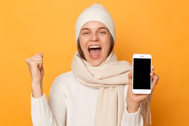 Retrato de una mujer positiva sonriente y emocionada que usa una bufanda de suéter y un sombrero que se encuentran aisladas sobre un fondo amarillo que muestra un teléfono celular con un espacio de copia vacío