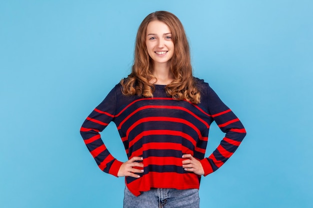 Retrato de una mujer positiva sonriente que usa un suéter de estilo casual a rayas mirando a la cámara manteniendo las manos en las caderas con una expresión segura