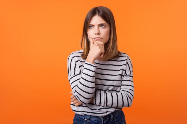 Retrato de una mujer preocupada y frustrada con el pelo castaño y un pantalones a rayas de manga larga de pie, sosteniendo su barbilla y frunciendo el ceño, pensando con una mirada seria. tiro de estudio interior aislado sobre fondo naranja