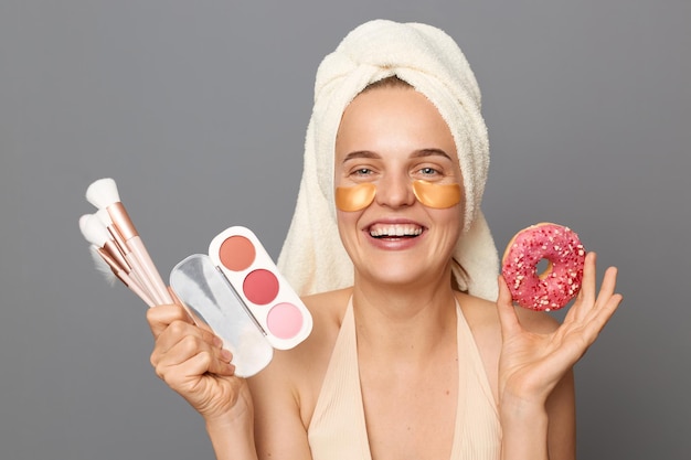 Retrato de una mujer que se ríe extremadamente feliz con una toalla en la cabeza sosteniendo sombras de ojos y pinceles para mostrar un delicioso donut mirando a la cámara de pie aislada sobre un fondo gris