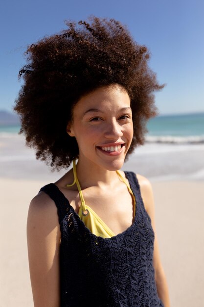 Retrato de una mujer de raza mixta con peinado afro, de vacaciones, disfrutando del tiempo libre sonriendo en una playa soleada, con cielo azul y mar de fondo