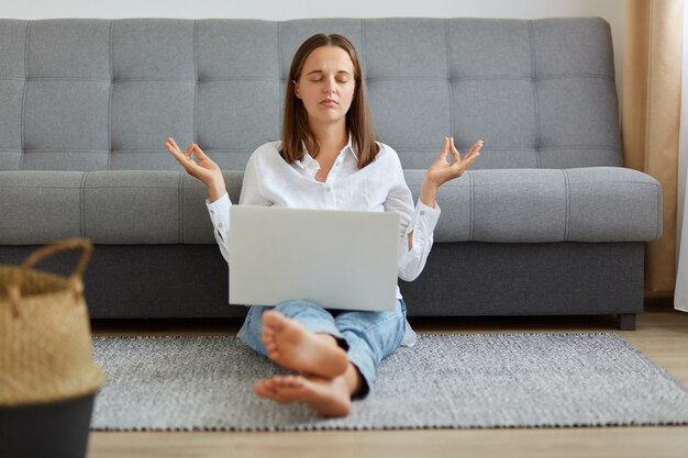 Retrato de mujer relajada con camisa blanca y jeans sentada en el piso con computadora portátil en las piernas, manteniendo las manos en gesto de yoga, tratando de calmarse y descansar.