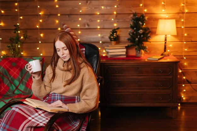 Retrato de una mujer relajada sosteniendo una taza con café caliente y leyendo un libro mientras se sienta en un sillón