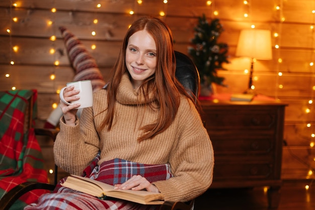 Retrato de una mujer relajada sosteniendo una taza con café caliente y leyendo un libro mientras se sienta en un sillón