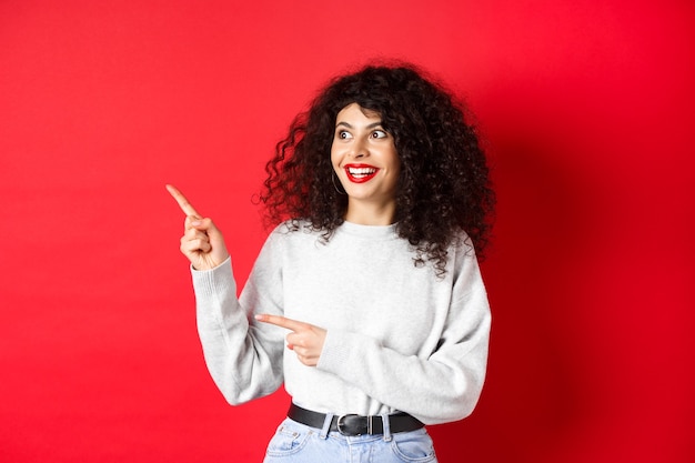 Retrato de mujer rizada emocionada con labios rojos, mirando y apuntando hacia la izquierda en el logotipo con cara de asombro, mirando la oferta promocional, de pie en la pared del estudio