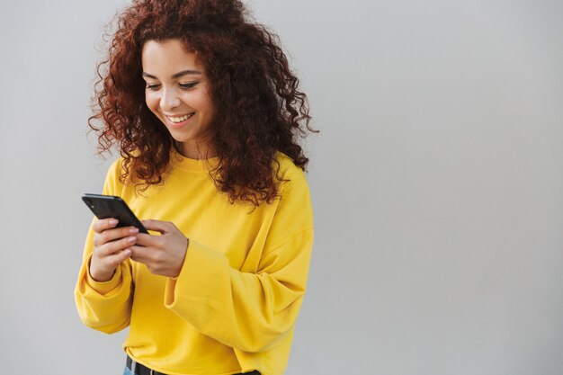 Foto retrato de mujer rizada hermosa alegre sonriente feliz aislada sobre pared gris mediante teléfono móvil.